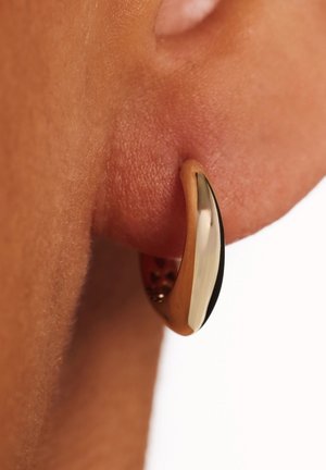 Gouden en zilveren oorring met een glanzende afwerking, met een gebogen ontwerp en een gestructureerd oppervlak, geklemd aan een oor.