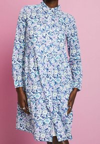 Femme portant une robe à manches longues et à boutons avec un motif floral bleu, violet et blanc sur un fond rose.
