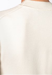 Zblízka pohled na zadní část ramene a krk osoby, která nosí krémově zbarvený žebrovaný pletený svetr.