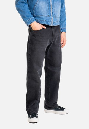 Person wearing black jeans, black and white sneakers, and a blue denim jacket with one hand in pocket against white background.