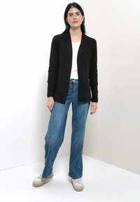 Cardigan noir en maille côtelée avec ouverture frontale, associé à un jean évasé bleu taille haute et des baskets blanches. Design simple et décontracté.