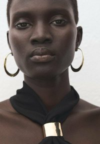 Gouden oorringen met hoepel vormen een set met een zwarte stoffen sjaal met een metalen gouden accent, die een close-up van een glad gezicht met neutrale tinten omlijst.