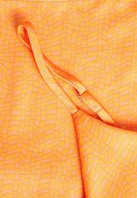 Pomarańczowa tkanina z teksturowanym, siatkowym wzorem. Zawiera detal w postaci paska przymocowanego do tkaniny, o gładkiej strukturze.