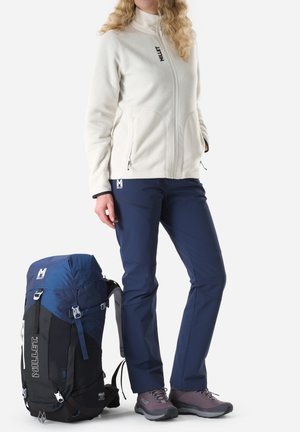 Woman wearing white fleece jacket and navy pants stands next to a large blue and black hiking backpack on a white background.