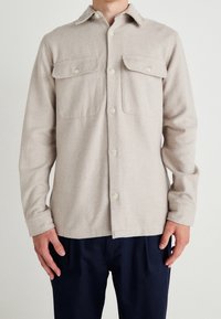 Jasnobeżowa koszula na guziki wykonana z teksturowanej tkaniny, z dwoma kieszeniami na piersi, kołnierzykiem i kremowymi guzikami. Noszona z ciemnymi spodniami.