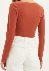 Maglia a maniche lunghe color ruggine in tessuto a costine con scollatura quadrata, abbinata a pantaloni bianchi a vita alta con tasche posteriori.