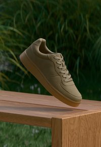 Μπεζ casual αθλητικό παπούτσι με κορδόνια να αιωρείται πάνω από ξύλινο παγκάκι, με θολό φόντο από πράσινα φύλλα.