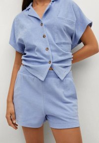 Mujer vestida con una camisa de botones de manga corta azul claro y pantalones cortos de cintura elástica a juego, de pie con una mano en la cadera frente a un fondo blanco.
