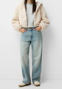 Felpa in pelliccia sintetica beige, con cappuccio, abbinata a jeans a gamba larga blu chiaro e sneakers bianche. Outfit casual con tessuti testurizzati.
