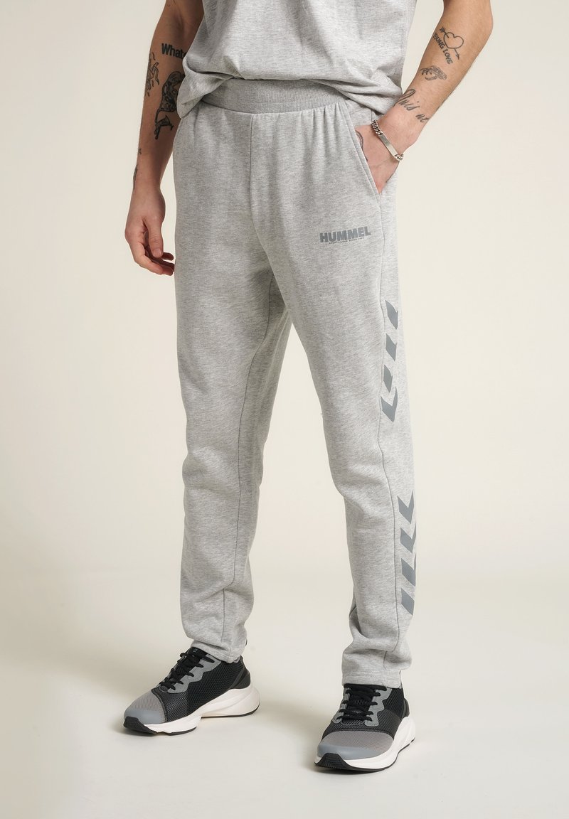 Grau Sweatpants mit einem Kordelzug-Bund, die ein Logo und ein Chevron-Muster in dunklerem Grau an den Seiten aufweisen. Kombiniert mit schwarzen und grauen Turnschuhen.