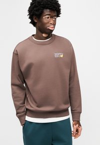 Καφέ φούτερ με ριμπ λαιμόκοψη και μανσέτες, με μικρό κεντημένο λογότυπο. Φοριέται πάνω από λευκό πουκάμισο με στρώσεις και συνδυάζεται με πετρόλ παντελόνια.