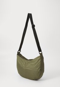 Olivno zelena torba preko ramena, izdelana iz lahke tkanine, s krožno obliko, nastavljivim črnim trakom in zadrgo.