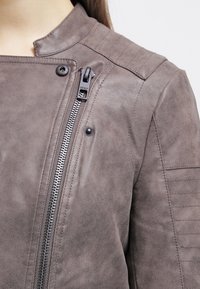 Braune Kunstlederjacke mit einem diagonalen Reißverschluss, Druckknopfdetails und genähten Akzenten an Schultern und Ärmeln. Glatte Textur.