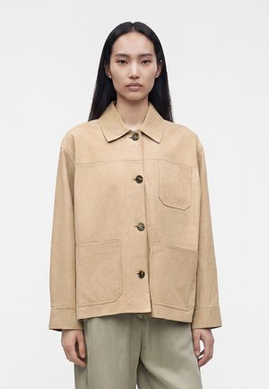 Jeune femme aux longs cheveux noirs portant une veste beige boutonnée avec trois grandes poches avant et un pantalon gris clair, faisant face à l'avant.
