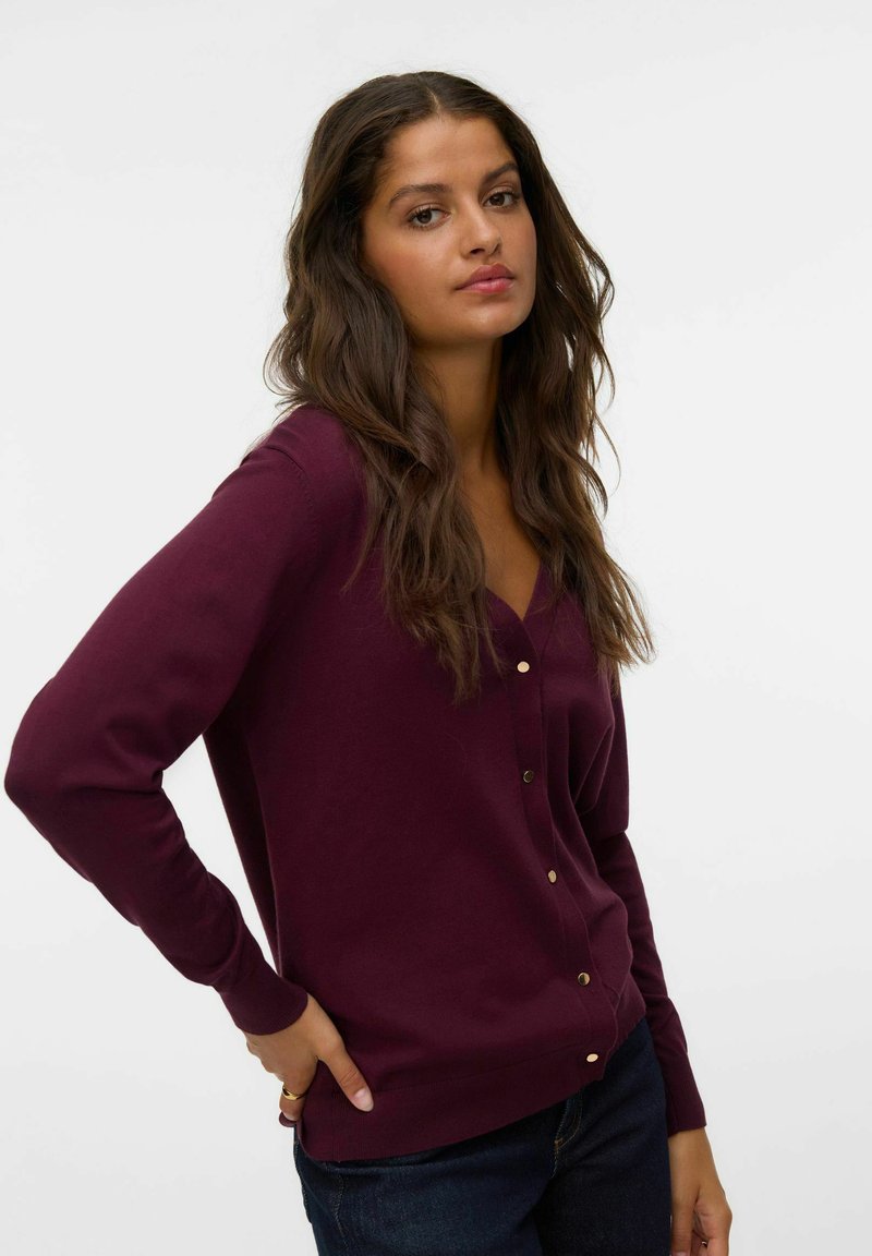 Cardigan bordeaux con scollo a V, maniche lunghe e bottoni dorati. Texture morbida e forma aderente, abbinato a jeans scuri.