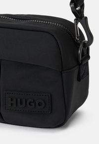 Piccola borsa a tracolla rettangolare nera con tasca frontale, chiusura a zip, clip per la tracolla e patch con il logo "HUGO" sulla parte anteriore.