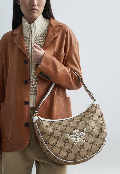 Nő barna bőr blézerben és bézs kötött pulóverben, aki egy barna mintás félhold táskát tart, fehér díszítéssel és MCM logóval.