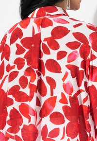 Motif de feuilles rouges et roses sur un tissu blanc, avec un col et un détail de pli au dos, présentant une texture lisse et légère.