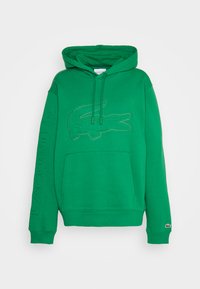 Sudadera verde con capucha hecha de una mezcla de algodón. Presenta un logotipo de cocodrilo bordado en la parte delantera y el texto "LACOSTE" en la manga.