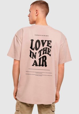 Kurze Ärmel pinkes T-Shirt mit einem schwarzen grafischen Druck auf der Rückseite, der "LOVE IN THE AIR" liest, sowie einem kleineren Text darüber. Glatte Baumwolloberfläche.