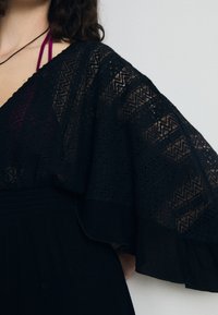 Svart spetsblus med vida ärmar som har ett zickzackmönster och genomskinlig textur. Elastisk midja med subtila lagerdetaljer.