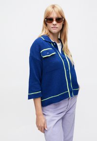 Niebieski dziergany kardigan z kołnierzem, neonowozielonymi akcentami i kieszeniami na guziki. W połączeniu z jasnofioletowymi sztruksowymi spodniami.