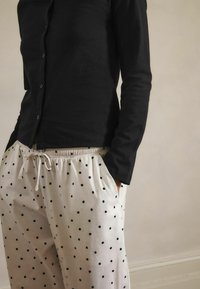 Person wearing a black long-sleeve top and white pants with black polka dots, hands in pockets, standing against a plain wall background.