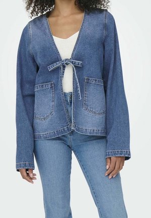 Vrouw draagt een medium blauwe spijkerjasje met voorstriksluiting en opgestikte zakken over een wit topje, gecombineerd met lichtblauwe jeans.