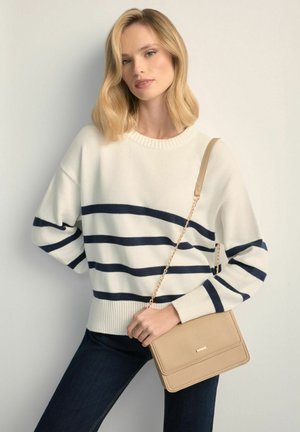 Blondynka w białym swetrze z granatowymi paskami, ciemnych dżinsach, trzymająca beżową torebkę z złotym łańcuszkiem.