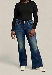 Personne portant un jean flare bleu foncé, un haut noir ajusté à manches longues et des baskets blanches, debout devant un fond uni.