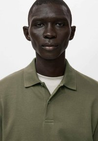 Tricou polo verde măsliniu cu guler clasic și fante cu nasturi, purtat peste un tricou alb cu gât rotund, având o textură fină.