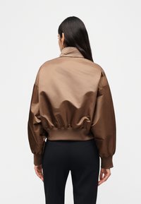 Γυναίκα με μακριά σκούρα μαλλιά που φοράει καφέ σατέν μπουφάν bomber και μαύρο παντελόνι, εμφανίζεται από πίσω σε απλό φόντο.