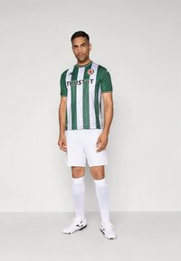 Futbalový dres so zelenými a bielymi pruhmi s logom „TRUST IT“ a hviezdičkovou nášivkou, doplnený bielymi šortkami a ponožkami a čierno-bielymi kopačkami.