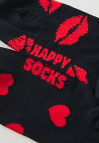 Chaussettes noires avec des motifs de cœurs rouges et de lèvres, avec le texte "HAPPY SOCKS" en rouge vif. Fabriquées en tissu doux et texturé.