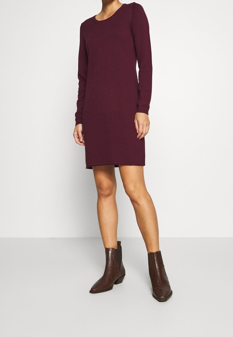 Femme portant une robe bordeaux à manches longues, longueur genoux, avec des bottines marron foncé, debout sur un sol gris clair.