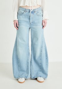 Jeans de tiro alto de mezclilla, de color azul claro y de pierna ancha, con grandes pliegues y un sutil desvanecimiento. Se ven zapatillas blancas en la parte inferior.