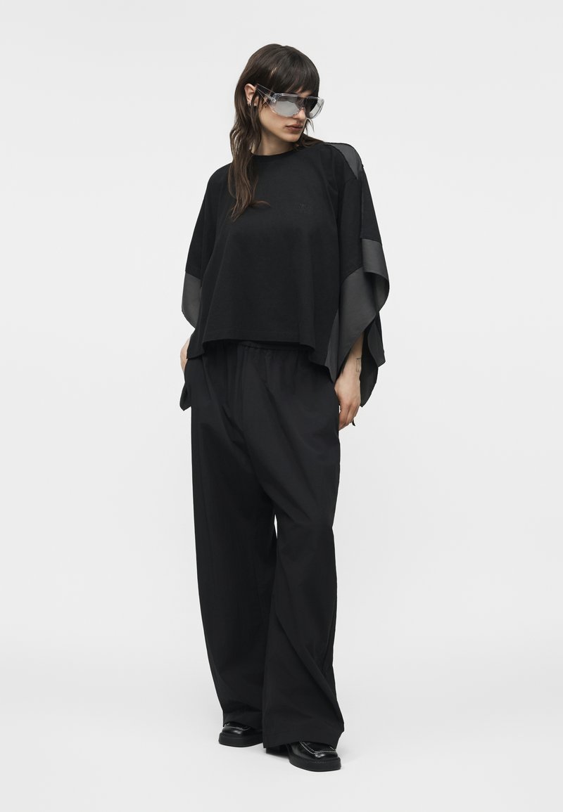 Femme portant un haut noir oversize avec des manches transparentes, un pantalon large noir, des chaussures noires et de grandes lunettes de soleil transparentes, debout devant un fond blanc.