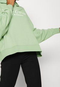 Felpa oversize verde chiaro con cappuccio regolabile e testo frontale. Presenta spalle ribassate e orlo curvo. Abbinata a pantaloni neri.
