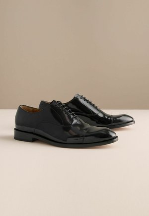 Czarne lakierowane pantofle z zaokrąglonym noskiem, zapinane na sznurowadła, z niewielkim obcasem i subtelnym przeszyciem wzdłuż szwów.