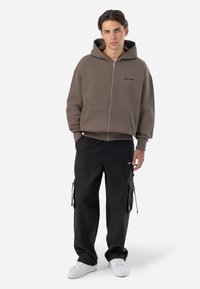 Brown zip-up hoodie with front pockets and logo. Paired with black cargo pants featuring side pockets and drawstring details. White sneakers.