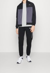 Chaqueta deportiva negra y gris con cremallera, que cuenta con un cuello alto y un logo. Combinada con pantalones negros que presentan paneles laterales en contraste. Zapatillas blancas.