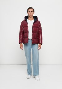 Man standing wearing a burgundy puffer jacket over a white shirt with light blue jeans and white sneakers on a plain white background.