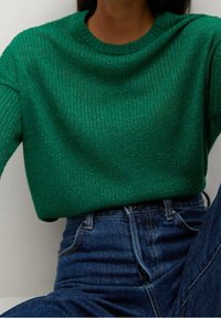 Vihreä ribbineulottu neule, joka on valmistettu rakenteellisesta materiaalista, jossa on pyöreä pääntie ja hieman löysä istuvuus, yhdistetty sinisiin farkkuihin.