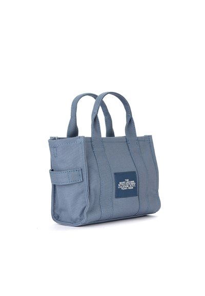 Saco de lona azul com laterais estruturadas, alças curtas e um bolso lateral. Apresenta um patch com o logotipo e fecho zippers na parte superior.