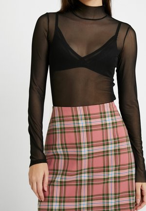 Femme portant un haut noir transparent à manches longues avec un bralette noir en dessous et une jupe à carreaux rose, verte et blanche.