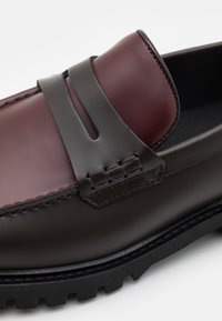 Mocassins en cuir marron foncé avec une bande supérieure en cuir bordeaux, présentant un détail découpé et des coutures contrastantes, sur une semelle noire épaisse.