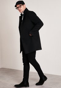 Black wool coat with a high collar, paired with fitted black trousers and black ankle boots. Glasses and a black cap add modern accents.