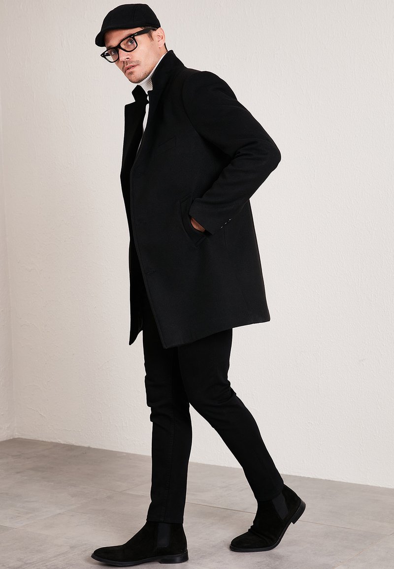 Black wool coat with a high collar, paired with fitted black trousers and black ankle boots. Glasses and a black cap add modern accents.