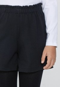 Czarne bawełniane szorty z elastycznym pasem, w luźnym kroju i miękkiej strukturze. W połączeniu z białą bluzką z długim rękawem i czarnymi rajstopami.