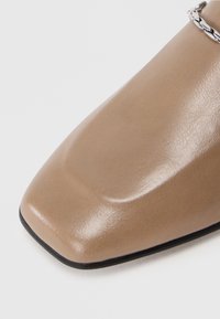 Beige lakkbőr cipő négyszögletes orral és sima felülettel, ezüst lánccal díszítve a felső részén.